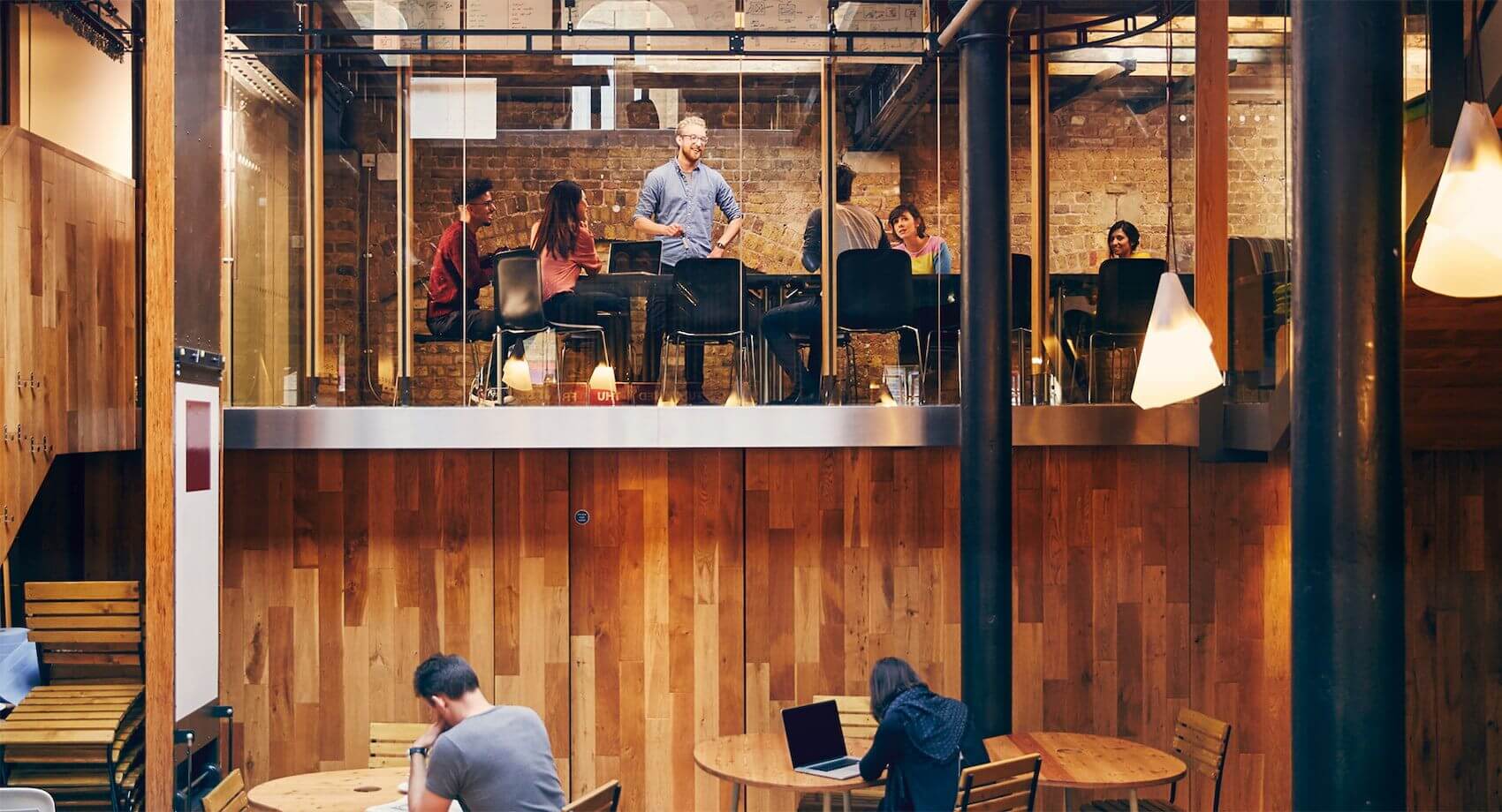 The photograph depicts a bustling office environment, with a group of people sitting at tables, engrossed in their work. The scene is characterized by an atmosphere of focus and productivity, with laptops and books strewn across the tables. The central focus of the composition is a group of people sitting around a large table, deep in discussion. A man in glasses stands in front of a brick wall, appearing to address the group. In the foreground, two individuals are seated at separate tables, typing away on their laptops. A white pendant light hangs over a kitchen counter in the background, casting a warm glow over the scene. Another table features a laptop and book placed side by side. The overall impression conveyed is one of a busy coffee shop or co-working space where people come together to collaborate and work on individual projects.