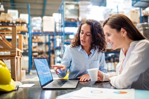 Young woman showing maintenance work order management on laptop to manager in factory warehouse. Two business women working together on laptop, checking the inventory.